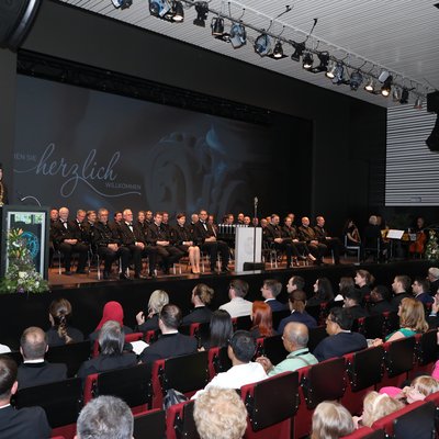 Ein großer Saal bei einer akademischen Feier: Publikum sitzt in Reihen, auf der Bühne eine Gruppe in festlicher Amtstracht, ein Redner am Pult, Beleuchtung von oben; Willkommensprojektion im Hintergrund.