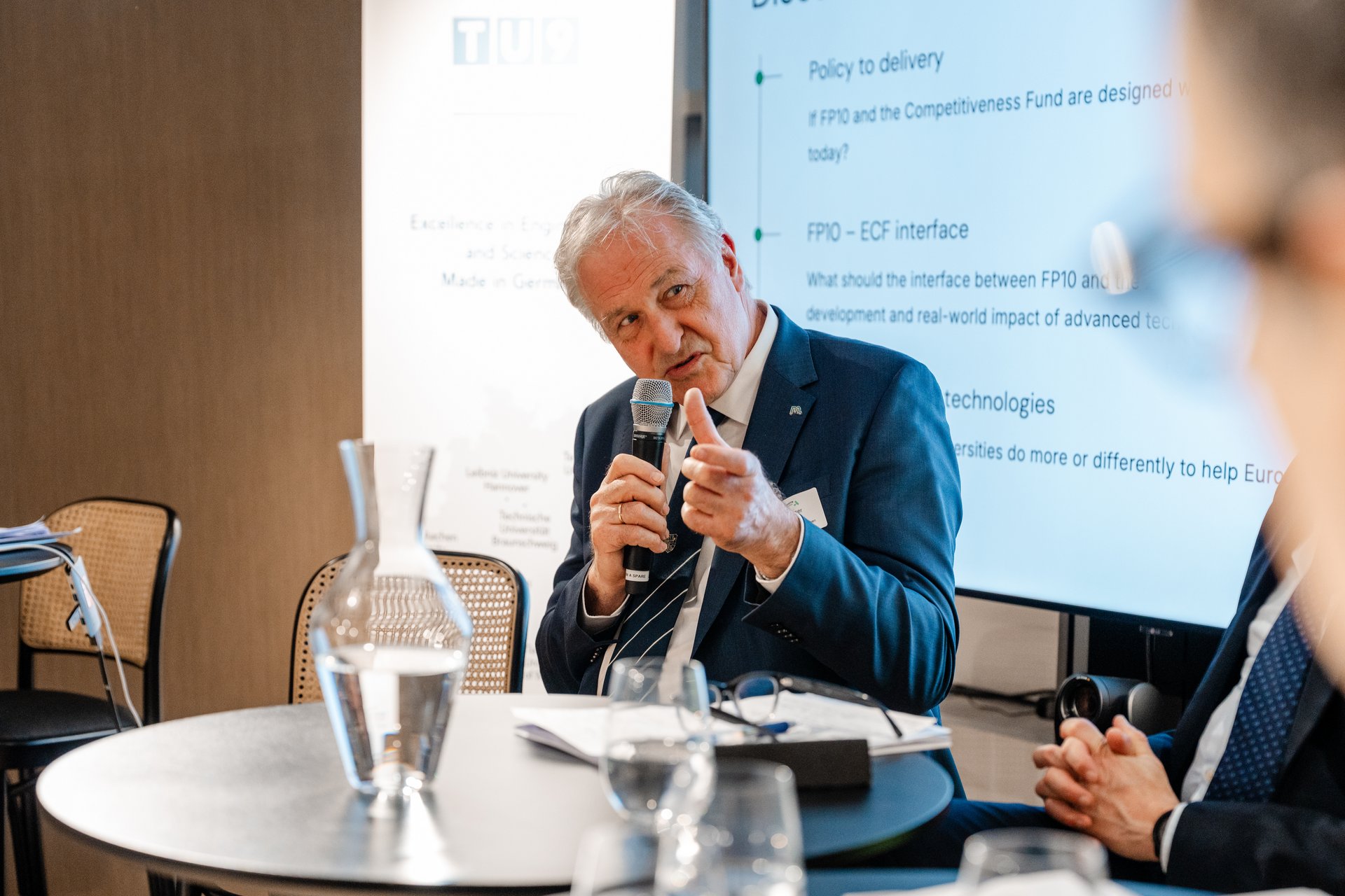 Rektor Peter Moser inmitten einer Podiumsdiskussion. Er hält ein Mikrofon in der Hand und zeigt einen Daumen nach oben.