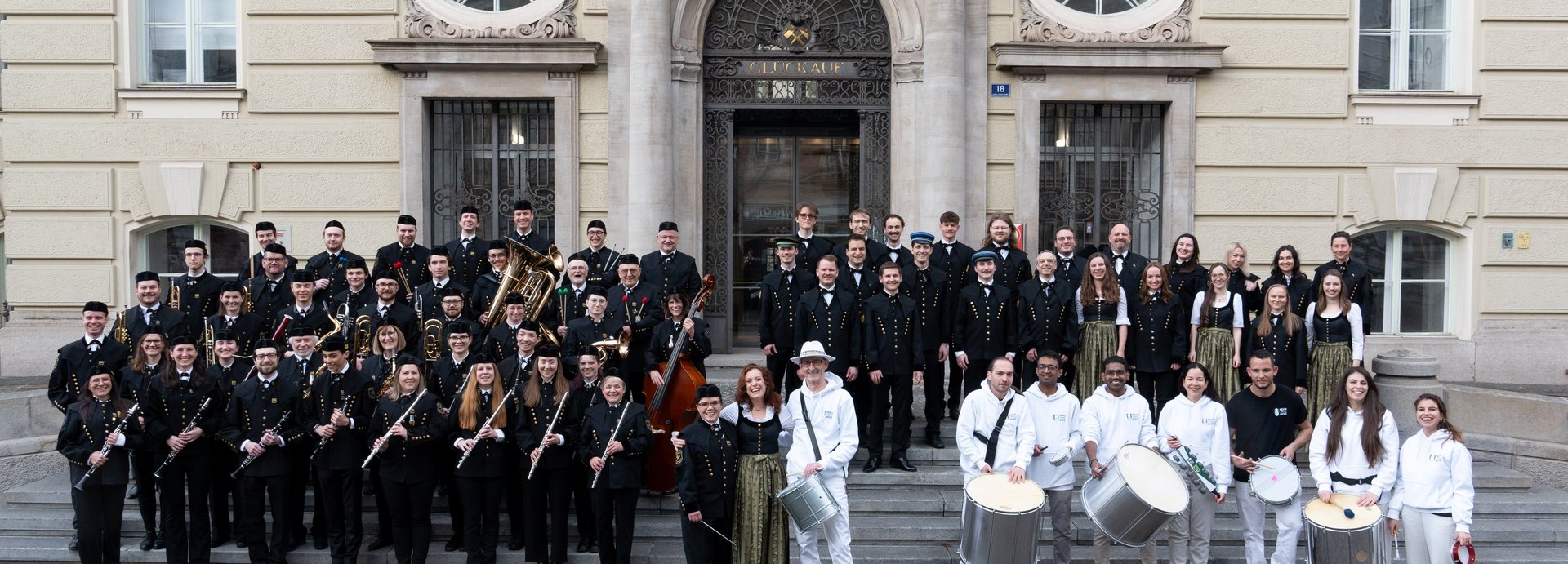 Die Mitglieder des Universitätsblasorchesters, des Universitätschors und die Trommelformation stehen zusammen auf den Außentreppen des Hauptgebäudes.