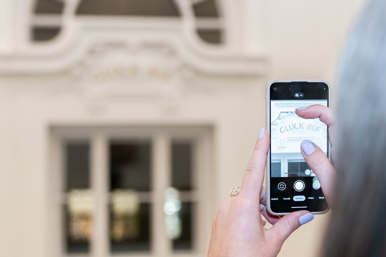 Verstärken Sie unseren Social Media Auftritt durch Ihre kreativen Beiträge! Foto: © MUL Eine Person fotografiert mit ihrem Smartphone den Leitspruch der Universität "Glück auf".