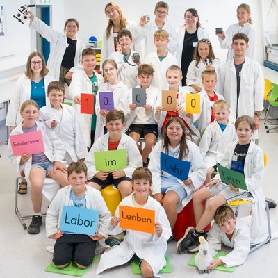 Schulklasse und Betreuer:innen in weißen Laborkitteln posieren im Labor und halten bunte Schilder: „10.000 Schüler*innen im Lehr‑Lern‑Labor Leoben".