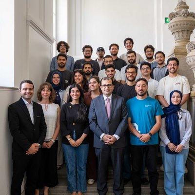 Eine große Gruppe Studierender und Universitätsangehöriger posiert auf einer Steintreppe mit verzierten Geländern in einem historischen Innenraum. Vorne stehen mehrere Personen in formeller Kleidung, dahinter weitere in legerer Kleidung.