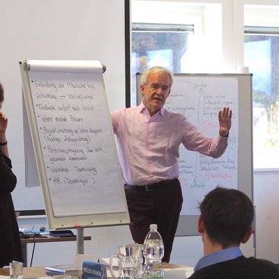 Ein Dozent der Delta Akademie an der Montanuniversität Leoben erklärt strategische Konzepte an einer Flipchart, während eine Gruppe von Studentinnen und Studenten aufmerksam zuhört und Notizen macht.