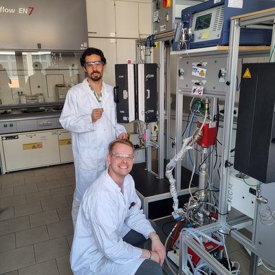 In einem Chemielabor stehen zwei Forschende in Laborkitteln und Schutzbrillen neben einer komplexen Reaktor‑/Messanlage mit Schläuchen, Sensoren und Steuergeräten.
