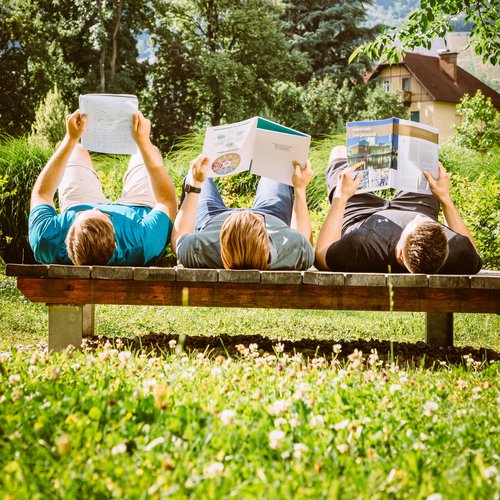 Drei Studenten liegen auf einer Bank in einer grünen Wiese und lesen Magazine der Montanuniversität.